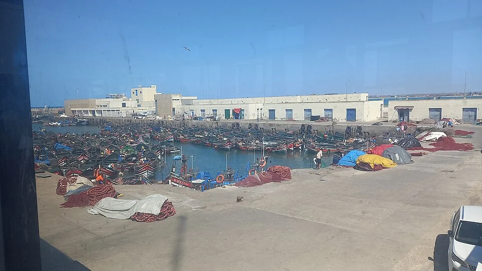 El Jadida balıkçı limanında küçük tekneler, kıyı yapıları ve malzemeler, 7 Mayıs 2025. Fotoğrafçı: Ahlamine; cam arkasından çekilmiş tarihli görüntü, restoran veya yolcu seferi değildir.