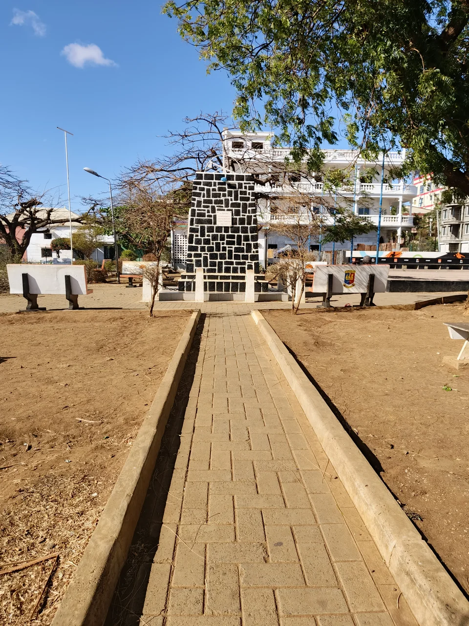 Path toward a stone monument, benches and trees at Place Kabary